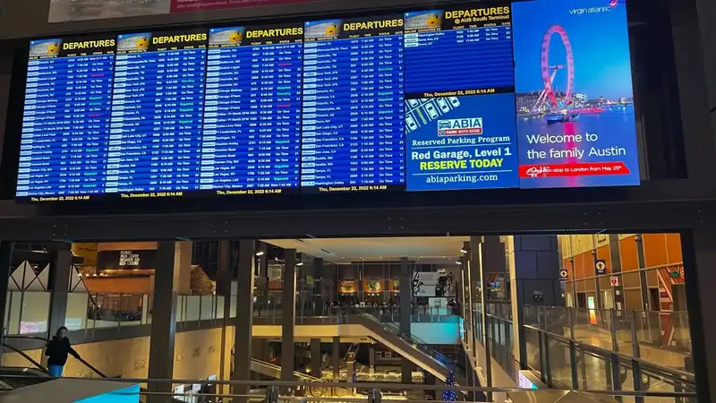 Interior of an airport showcasing large flight departure screens with schedules, an advertisement for parking at ABIA, and a welcoming digital billboard saying 'Welcome to the family Austin' with an image of a Ferris wheel.