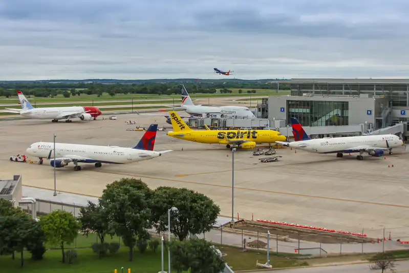 Aerial view of Austin-Bergstrom International Airport with various commercial airplanes at the gates, including a prominently featured Spirit Airlines jet, and another airplane mid-takeoff in the background.