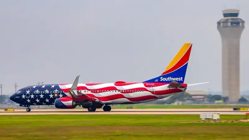 A Southwest Airlines airplane adorned with an American flag-themed livery takes off from a runway, with the control tower of Austin-Bergstrom International Airport visible in the background